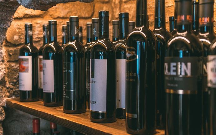A row of wine bottles on a wooden shelf. The bottles are in dark tones and placed against a stone wall.