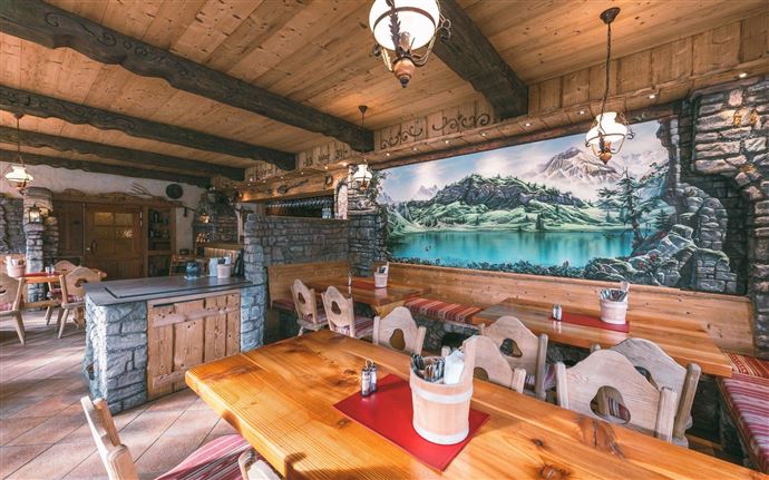 A rustic restaurant with wooden decor and warm lighting. On the wall, there is a large painting of a mountain landscape.