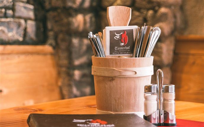 A wooden pot with cutlery and a menu is placed on a table. The surroundings appear rustic and inviting.