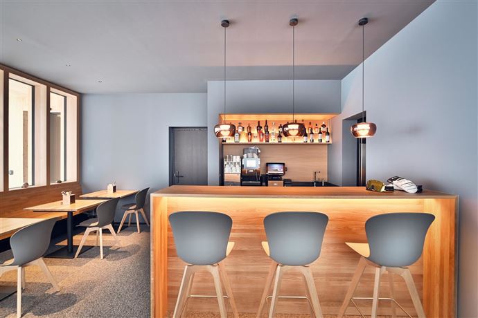 Eine moderne Bar mit Holzregal und eleganten Stühlen. Helle Wände und gemütliche Atmosphäre schaffen einen einladenden Raum.