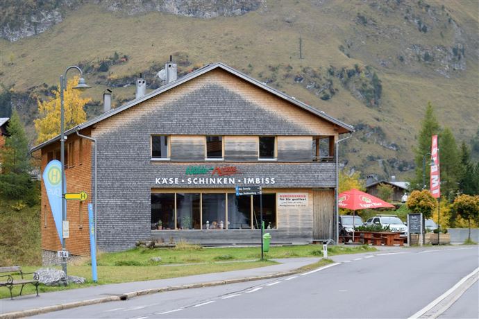 A rustic building with a snack bar that offers cheese and ham. There are seating areas outside and the surrounding landscape is mountainous.