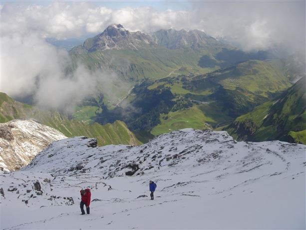 Twee wandelaars lopen over met sneeuw bedekte bergen met een groene dal landschappen op de achtergrond. De wolken trekken over de hoge toppen.