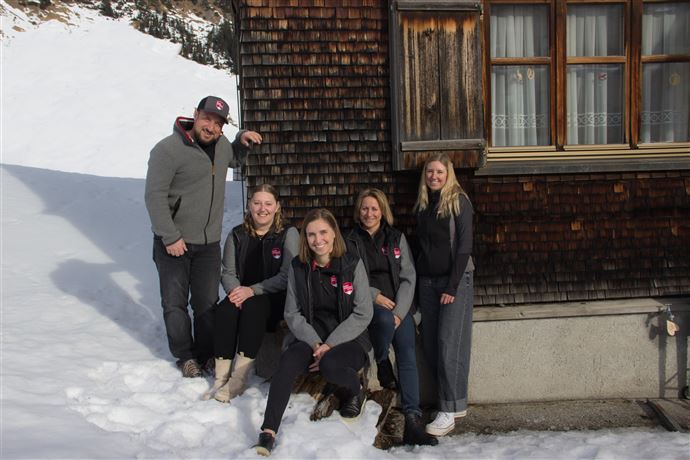 A group of five people poses in front of a wooden house in the snow. In the background, snow-covered landscapes can be seen.