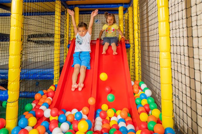 Two children happily slide down red slides into a pool of colorful balls. The surroundings are fresh and designed to be child-friendly.