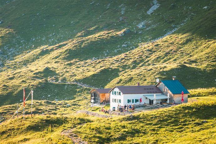 A mountain cabin surrounded by green meadows and gentle hills. People are gathering in front of the building in nature.