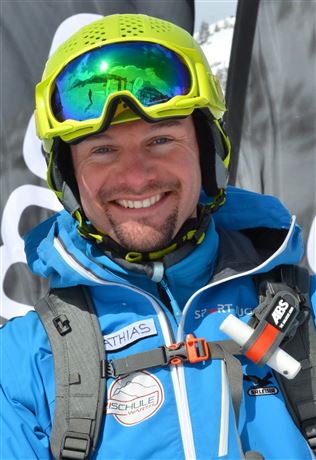 A laughing man in a blue ski outfit with a yellow helmet and reflective sunglasses. He is standing in front of a snowy background.