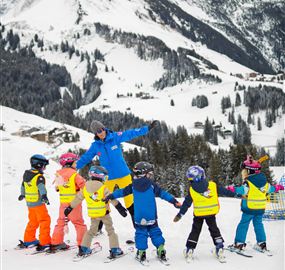 A group of children in colorful ski suits stands in the snow and learns to ski. In the background, snow-covered mountains and a clear sky can be seen.
