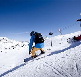 Two snowboarders are riding down the snow in a sunny ski resort. In the background, snow-covered mountains can be seen.
