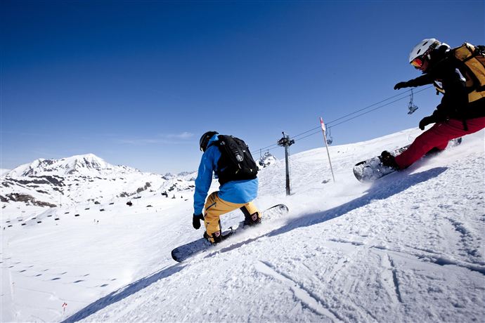 Zwei Snowboarder fahren den Schnee in einem sonnigen Skigebiet hinunter. Im Hintergrund sind schneebedeckte Berge zu sehen.