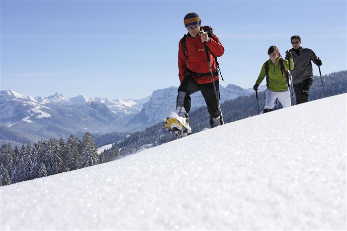 Drei Personen wandern durch verschneite Landschaften in den Bergen. Der Himmel ist klar und blauer als ein Wintertag.