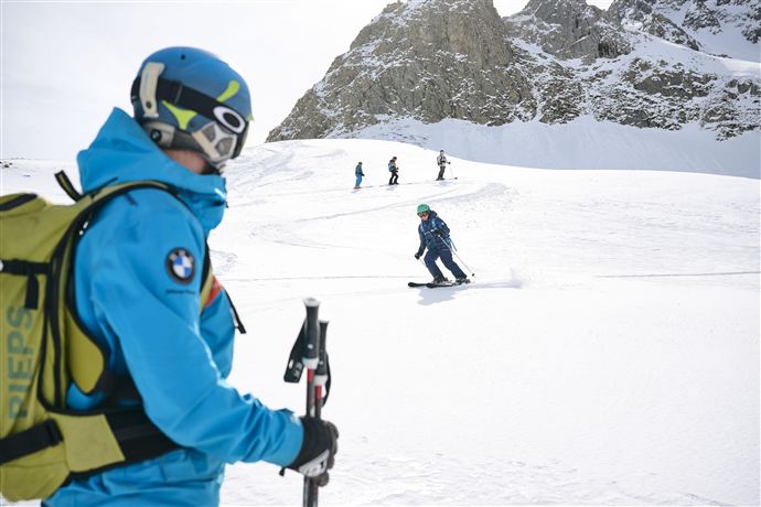 Ein Skilehrer in blauer Ausrüstung beobachtet Schüler, die den Hang hinunterfahren. Die schneebedeckte Landschaft und die Berge im Hintergrund schaffen eine winterliche Atmosphäre.