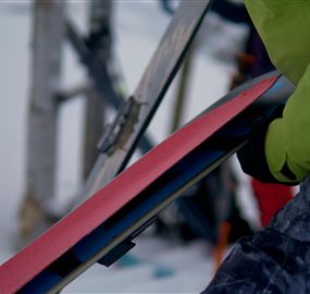A person is holding a red snowboard in their hand. In the background, parts of a ski lift and snowy landscape can be seen.