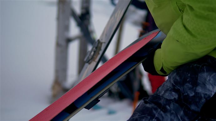 Eine Person hält ein rotes Snowboard in der Hand. Im Hintergrund sind Teile eines Skilifts und verschneite Landschaft zu sehen.
