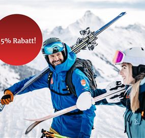 Two skiers are smiling and carrying their ski equipment in a snow-covered mountain landscape. In the background, a sign indicating a 15% discount is visible.