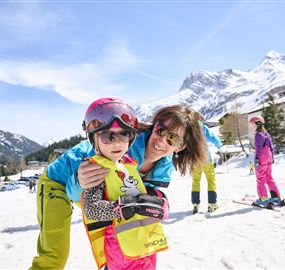 A happy child with a helmet and ski goggles stands in the snow, embraced by an adult. In the background, snowy mountains and other skiers can be seen.
