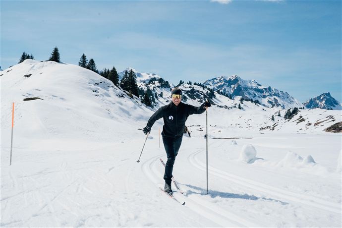 A person skiing stands on a snow-covered slope. In the background, snow-covered mountains and a clear sky can be seen.