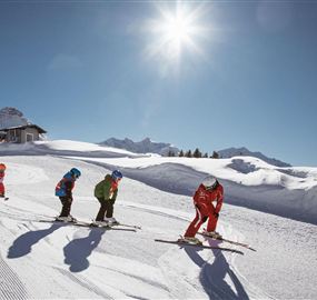 A group of skiers is skiing on a closed slope in the sun. In the background, snow-covered mountains and a building are visible.