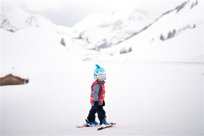 Ein kleines Kind fährt Ski in einer schneebedeckten Landschaft. Die Berge im Hintergrund sind ebenfalls mit Schnee bedeckt und der Himmel ist bewölkt.