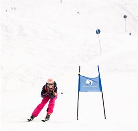 A skier in pink gear is skiing through a flag pole course in the snow. The background shows a snowy landscape with other skiers.