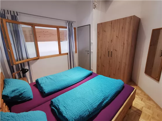 A cozy bedroom with two single beds in vibrant colors. The room has a window with curtains and a wooden closet against the wall.