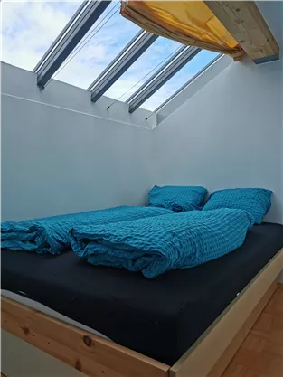 A cozy bedroom with a double bed. The bedding is in light blue, and large windows let in plenty of light.
