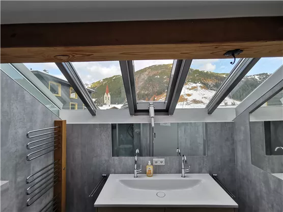 A modern bathroom with a large sink and a skylight. In the background, snow-covered mountains and a church are visible.