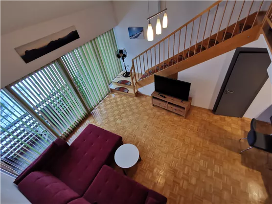 A modern living environment with a red sofa and a round table. Large windows let in plenty of light and provide a view of a wooden staircase.