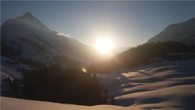 A winter mountain landscape with snow-covered hills. The sun rises behind the mountains and illuminates the scene.