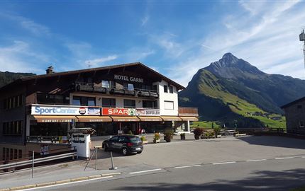 Ein Hotelgebäude mit dem Namen "Hotel Garni" in einer landschaftlichen Umgebung. Im Hintergrund sind Berge und ein blauer Himmel zu sehen.