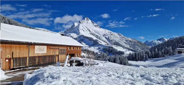 A picturesque winter landscape with snow-covered mountains and a wooden house. The sky is clear and blue, creating a peaceful atmosphere.