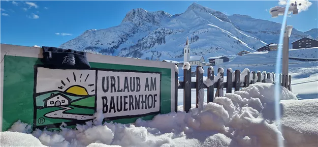 A sign with the inscription "Holiday on the Farm" stands in the snow. In the background, snow-covered mountains and a blue sky can be seen.