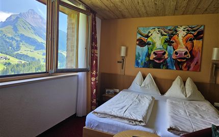 Ein gemütliches Hotelzimmer mit zwei Betten und einer farbenfrohen Kuhmalerei an der Wand. Durch das Fenster ist eine beeindruckende Berglandschaft zu sehen.