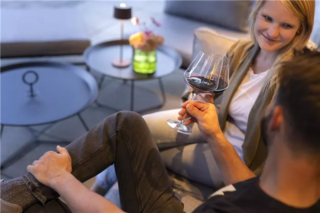 A relaxed atmosphere with a couple sitting together and enjoying wine. In the background, stylish furniture and a blooming vase can be seen.