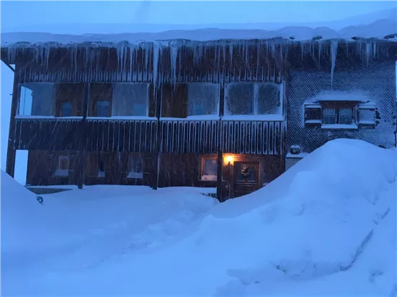 Ein Holzgebäude, das in tiefem Schnee verborgen ist, mit Eiszapfen an den Fenstern. Das Licht aus einem Fenster sorgt für eine gemütliche Atmosphäre in der winterlichen Szene.
