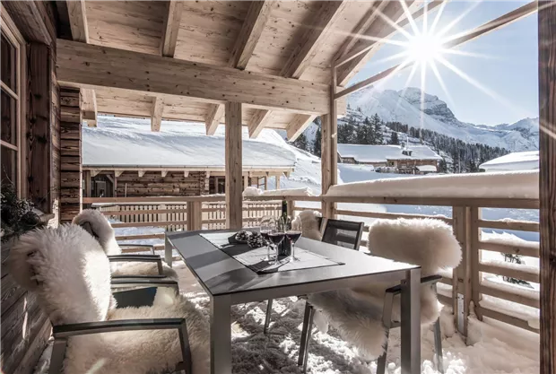 Eine gemütliche Terrasse mit einem Tisch und weichen Stühlen, umgeben von schneebedeckten Bergen. Die Sonne strahlt hell und schafft eine einladende Atmosphäre.