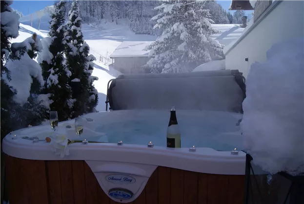 A relaxing hot tub in the snow with a bottle of champagne and glasses. In the background, there are snow-covered trees and a tranquil, wintry landscape.