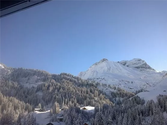 A snow-covered mountain landscape under a clear blue sky. The trees are covered with snow, and there are some small huts in the foreground.