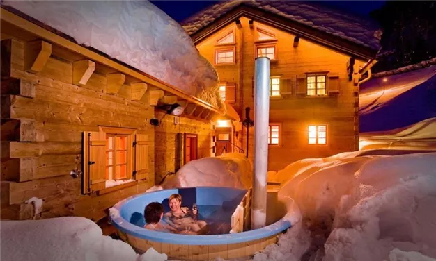 Ein Whirlpool im Freien, umgeben von Schnee und einem gemütlichen Holzhaus. Die warmen Lichter des Hauses schaffen eine einladende Atmosphäre in der Winterlandschaft.