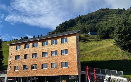 Ein modernes Gebäude mit Holzfassade, umgeben von grünen Hügeln. Der Himmel ist teilweise bewölkt und es gibt sichtbare Bäume.