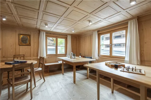 A bright room with wooden walls and tables. There are windows with a view outside and some chairs, as well as a chess game on the table.