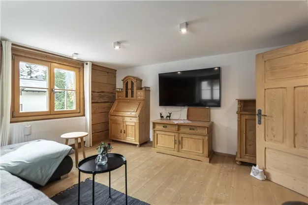 A cozy room with wooden furniture and a TV. Large windows let in a lot of light and create an inviting atmosphere.