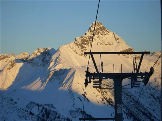 Eine schneebedeckte Berglandschaft mit einem klaren blauen Himmel. Im Vordergrund ist eine Skiliftgondel zu sehen.