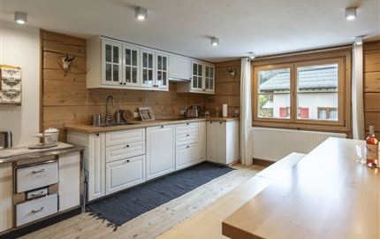 Eine moderne Küche mit Holzverkleidung und weißen Schränken. Große Fenster bieten viel Tageslicht und einen Blick auf die Außenlandschaft.