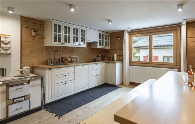 A modern kitchen with wooden paneling and white cabinets. Large windows provide plenty of daylight and a view of the outdoor landscape.