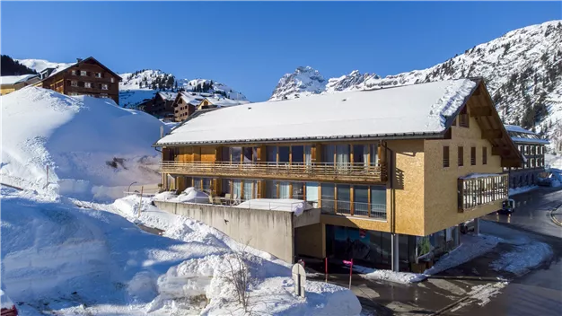 A cozy wooden building in the snow with a stunning mountain landscape in the background. The sky is blue and clear.