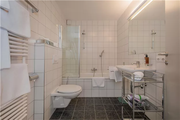 A modern bathroom with white tiles and a bathtub. There is a shower, a sink, and towels and toiletries.