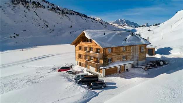 A cozy wooden house in a snowy landscape. In the background, mountains and a blue sky can be seen.