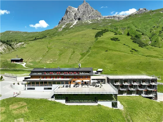 A modern hotel is located amidst a green mountain landscape. In the background, majestic mountains and a clear blue sky can be seen.