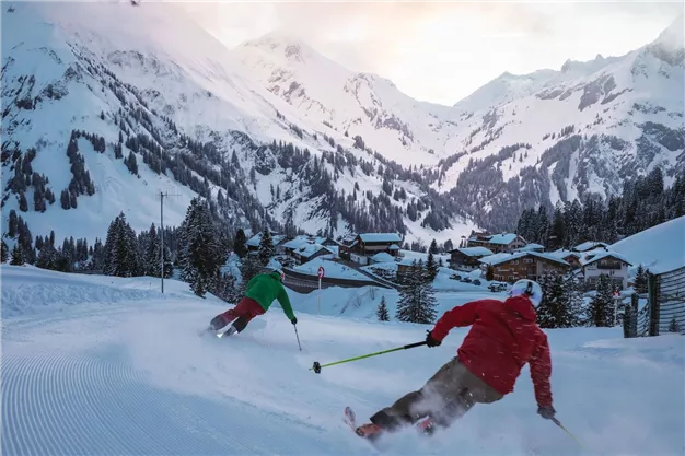 Zwei Skifahrer fahren eine schneebedeckte Piste hinunter. Im Hintergrund sind Berge und eine malerische alpine Landschaft zu sehen.