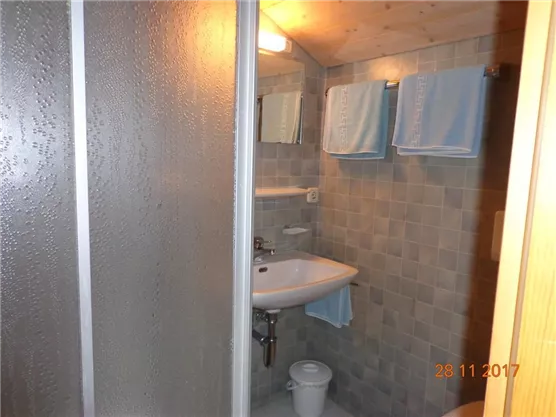 Ein kleines Badezimmer mit einer Dusche, einem Waschbecken und einer Toilette. Die Wände sind mit hellen Fliesen ausgekleidet, und es hängen blaue Handtücher.